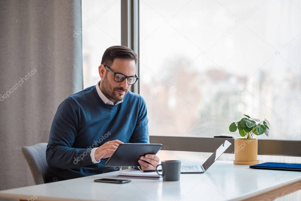 Young businessman using digital tablet while working on laptop. Male professional with wireless computer at desk. He is sitting by window in office.