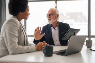 Dijital tablet üzerinden kadın partnerle yaşlı profesyonel planlama stratejisi. İş arkadaşları masada tartışıyor. Şirket ofisinde oturuyorlar..