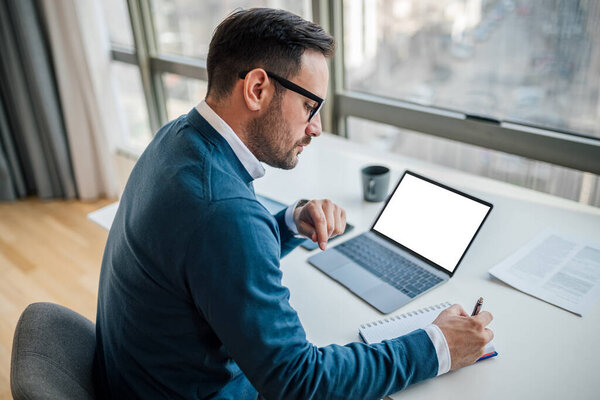 High angle view of serious businessman writing notes in diary. Male executive is using laptop while working at desk. White blank clean empty mock up template background computer laptop screen.