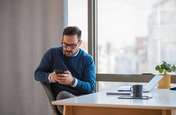 Young male entrepreneur messaging online on mobile phone. Businessman working on laptop at desk. He is wearing casuals in corporate office.