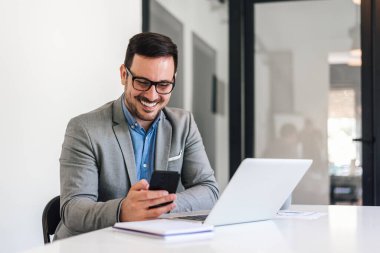 Genç gülümseyen iş adamları internet üzerinden akıllı telefonlardan iletişim kuruyor. Formalitelerdeki mutlu erkek profesyonel dizüstü bilgisayar üzerinde çalışıyor. Ofisteki masada oturuyor..