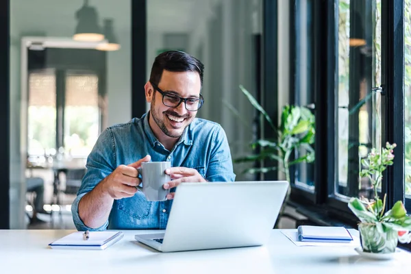 Happy male professional having coffee. Young smiling businessman looking at successful business plan on laptop. He is working at table by window in his home or office.