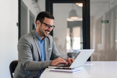 Dizüstü bilgisayarla çalışan neşeli genç bir profesyonel. Gülümseyen iş adamı başarılı iş planını analiz ediyor. Şirket ofisindeki masada oturuyor..
