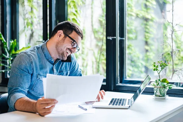 Happy professional discussing on smart phone while using laptop. Young male entrepreneur with documents multitasking at table. He is wearing casuals while working at home.