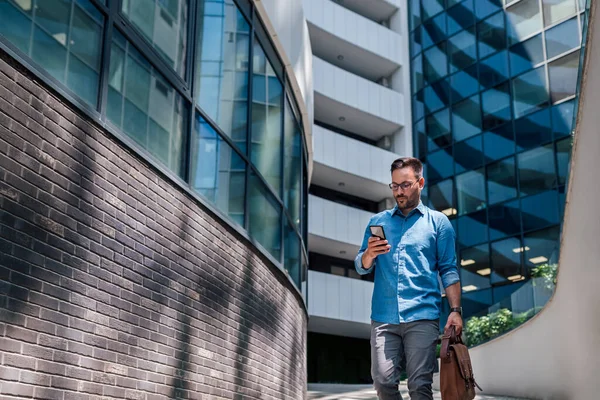 Confident professional networking on smart phone. Young businessman wearing formals is carrying laptop bag. He is walking on footpath against office building.