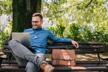 Laptop kullanan kendine güvenen bir iş adamı. Erkek profesyonel kablosuz teknoloji üzerinde çalışıyor. Kamu parkında bir çantayla oturuyor..