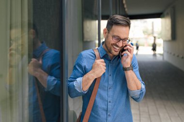 Gülümseyen bir profesyonel cep telefonuyla konuşuyor. Genç iş adamı dizüstü bilgisayar çantası taşıyor. Şehirdeki ofis binasının camını yansıtıyor..
