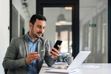 Akıllı telefondan kendine güvenen erkek yöneticilerin yakın çekim görüntüleri. Gözlüklü genç girişimci cep telefonundan mesaj okuyor. Ofis masasında dizüstü bilgisayarla çalışıyor..
