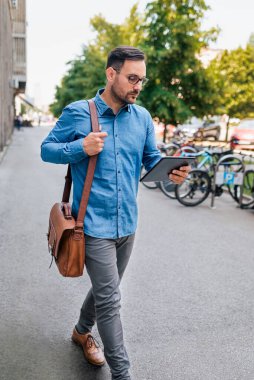 Ciddi bir iş adamı dijital tablet üzerinde analiz planı yapıyor. Profesyonel bir banliyö yolcusu dizüstü bilgisayar çantası taşıyor. O, şehirde kaldırımda yürüyor..
