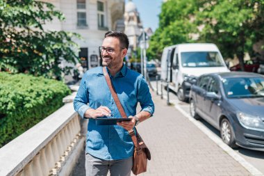 Dijital tablet kullanarak gülümseyen girişimci. Erkek yönetici kaldırımda yürürken gözünü kaçırıyor. Güneşli bir günde şehirde resmi kıyafet giyiyor..