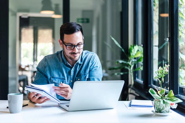 Happy professional analyzing business ideas in diary at table. Businessman making success plan while working on laptop. He is wearing casuals in home office or business office.