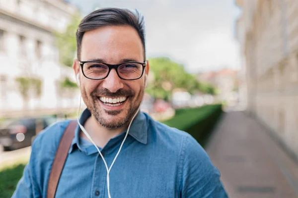Smiling young businessman using headphones. Portrait of male professional commuter listening music. He is wearing eyeglasses and formals in the city.