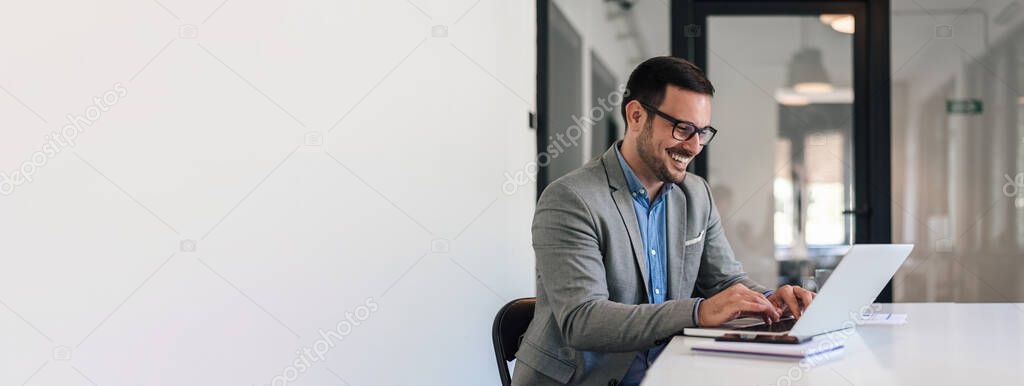 Cheerful young professional working on laptop. Smiling businessman is analyzing successful business plan. He is sitting at desk in corporate office.