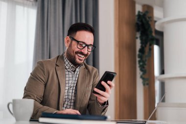 Resmi giysiler içinde gülümseyen bir işadamı cep telefonu kullanarak oturmuş elinde akıllı telefon tutarak kahve içiyor. Kahve molası verirken cep telefonuna bakan küçük işletme girişimcisi .