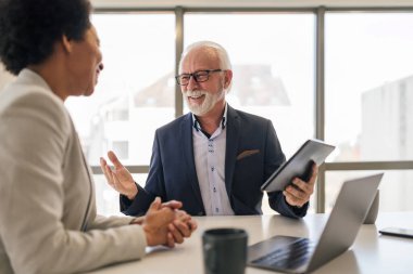 Elderly professional planning strategy with female partner over digital tablet. Business colleagues are discussing at office desk. They are sitting in corporate office.
