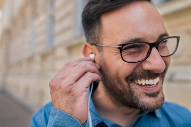 Smiling young businessman listening music on headphones. Male professional commuter is standing on sidewalk. He is enjoying while commuting in the city.