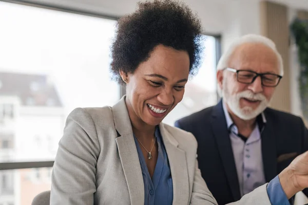 Multiracial woman laughing at funny joke with diverse senior male coworker in office. Friendly work team looking at documents and enjoying positive emotions together. Happy colleagues concept