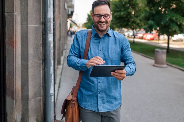 Smiling entrepreneur using digital tablet. Male executive commuter is looking at the screen of hand held digital tablet while walking on sidewalk. He is wearing formals in the city during sunny day.