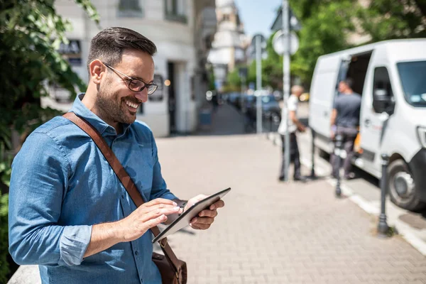 Portrait of happy entrepreneur holding digital tablet and looking at the screen. Young handsome businessman carrying laptop bag. He is standing on sidewalk in the city.