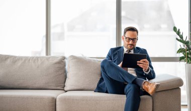 Ofiste dijital tablet kullanan kendinden emin bir erkek girişimci. İş adamı pencereye karşı kanepede oturuyor. İş yerindeki kablosuz bilgisayarda çalışıyor..