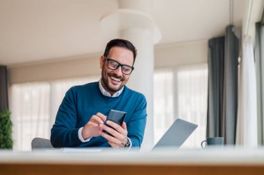 Gülümseyen iş adamı evde ya da ofiste telefon kullanıyor. Küçük işletme girişimcisi cep telefonuna bakıyor ve gülümsüyor ya da iyi haberler yolluyor..