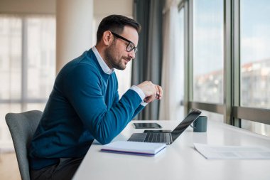 Masada otururken dizüstü bilgisayarda çalışan gülümseyen genç girişimci. Kendine güvenen profesyonel başarılı stratejiyi analiz ediyor. Ofiste çok şık giyiniyor..