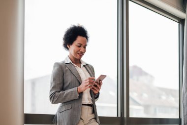 Gülümseyen bayan yönetici akıllı telefondan mesaj atıyor. İş kadını pencerenin önünde duruyor. Ofiste forma giyiyor..
