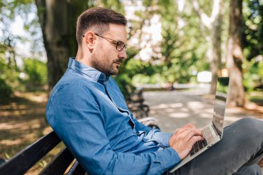 Serious male entrepreneur using laptop. Confident male executive is analyzing business plan on wireless computer. He is sitting on bench at the public park.