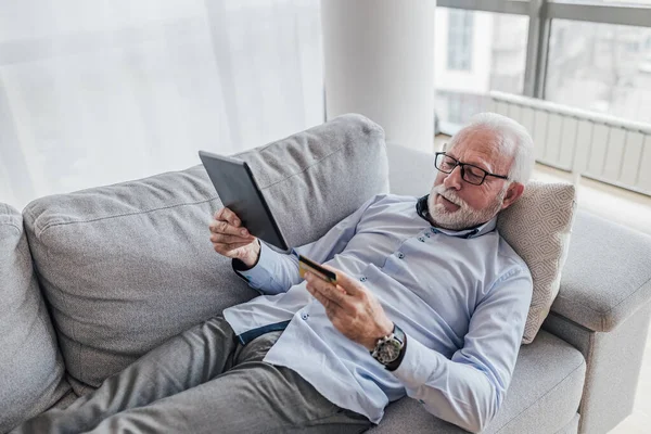 Thoughtful senior executive analyzing digital tablet. Freelancer making online payment through credit card. He is lying on sofa at home office.