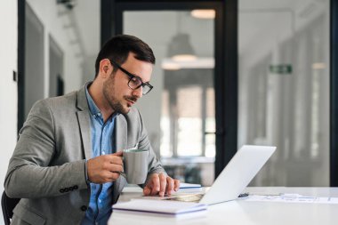 Serious male manager having coffee while working on laptop. Young businessman wearing eyeglasses is sitting at desk. He is working in corporate office.
