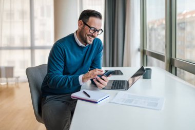 Cheerful entrepreneur networking on smart phone. Young male professional is working on laptop at desk. He is wearing business casuals in office.