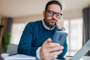 Close-up of bored executive text messaging on mobile phone. Young businessman is working at desk in office. He is wearing smart casuals at workplace.