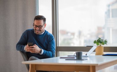 Smiling entrepreneur socializing online using smart phone. Young businessman is distracted while working at desk. He is sitting in office.