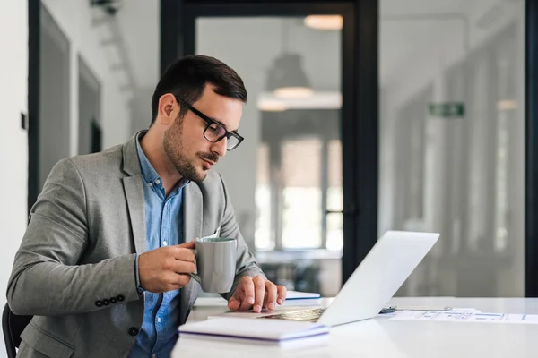 Serious male manager having coffee while working on laptop. Young businessman wearing eyeglasses is sitting at desk. He is working in corporate office.