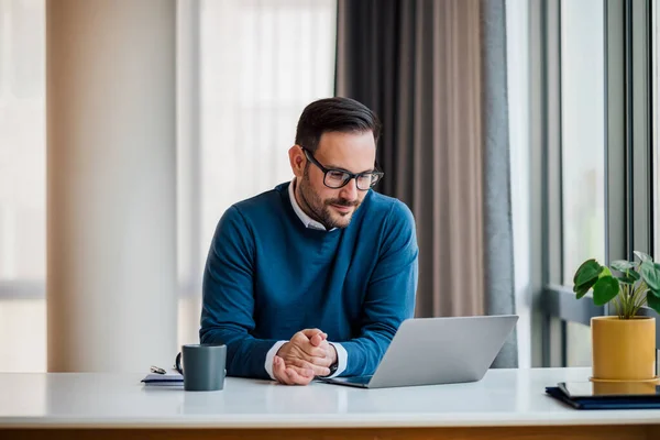Confident smiling young professional looking at successful business plan on laptop. Male manager is sitting with hands clasped at desk. He is working in office.