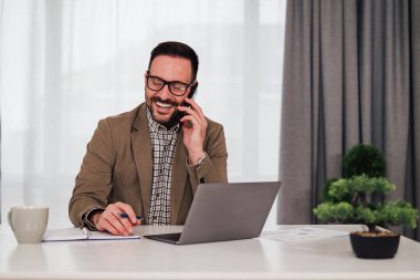 Cheerful male manager talking on smart phone. Young professional working on laptop at desk. He is wearing eyeglasses in the office.