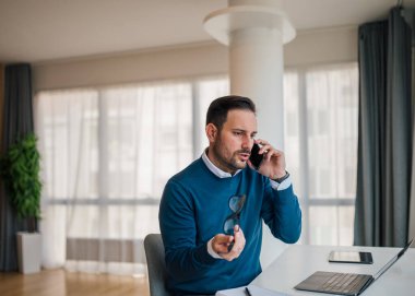 Stressed entrepreneur talking on smart phone. Angry male professional is working on laptop at desk. He is wearing smart casuals in office.