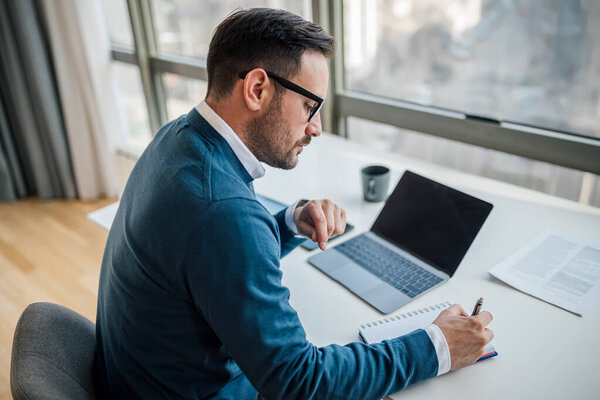 High angle view of serious professional writing notes in diary. Young businessman is using laptop while working at desk. He is wearing sweater in corporate office.