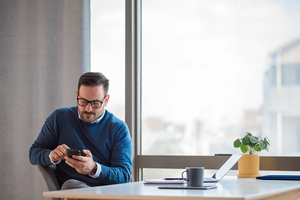 Young male professional messaging online on smart phone. Distracted businessman networking while working on laptop at desk. He is wearing casuals in corporate office.