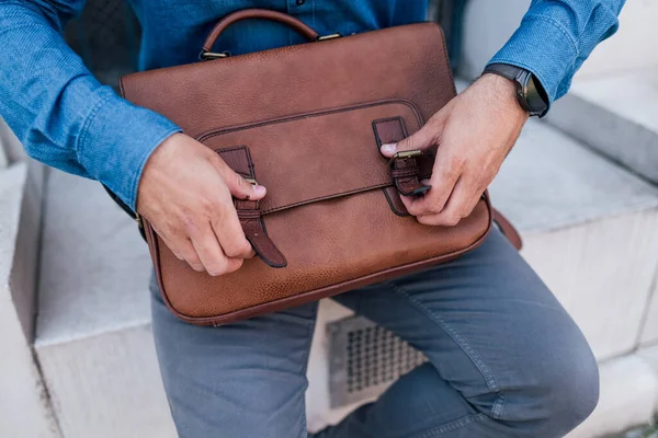 Midsection of businessman closing laptop bag. Male professional is wearing formals. He is sitting against building in the city.