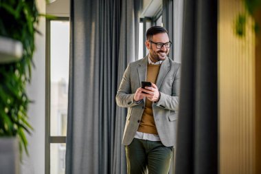 Calm adult man, looking through the window, while using his phone, leaning on window.