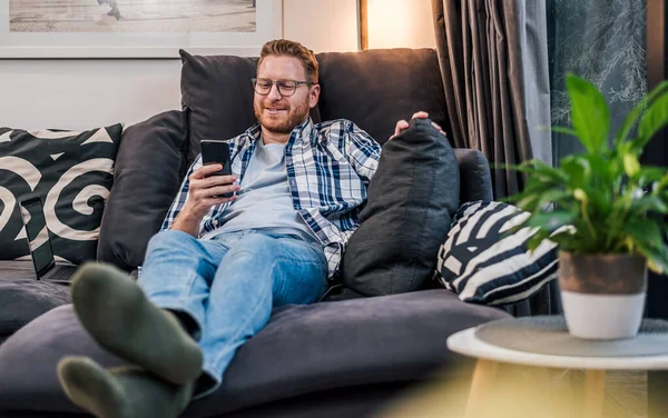 Delighted adult man laying on the couch, enjoying his free time using his mobile phone.