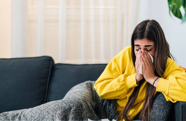 Fatigued young brunette woman, laying sick at home on the couch, sneezing.