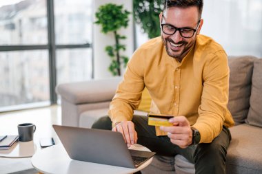 Gözlüklü, mutlu ve yetişkin bir adam. Evde, kredi kartından numarayı okuyor ve gülümsüyor..