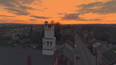 A Drone Approaching a View of a Small Town and a Steeple at Sunrise with Orange and Reds on a Spring Sunrise With Partial Cloudy Skies.