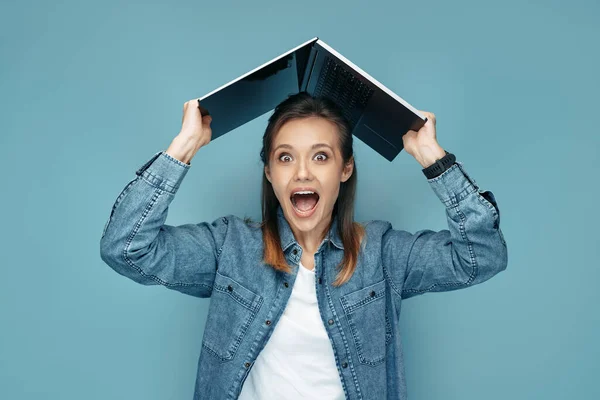 Opgewonden jonge vrouw in jeans shirt met computer of laptop over hoofd in de voorkant van de blauwe achtergrond. — Stockfoto