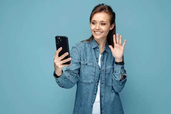 Cheerful young woman chatting on smartphone over blue background. Blogger. Waving hand. — 스톡 사진