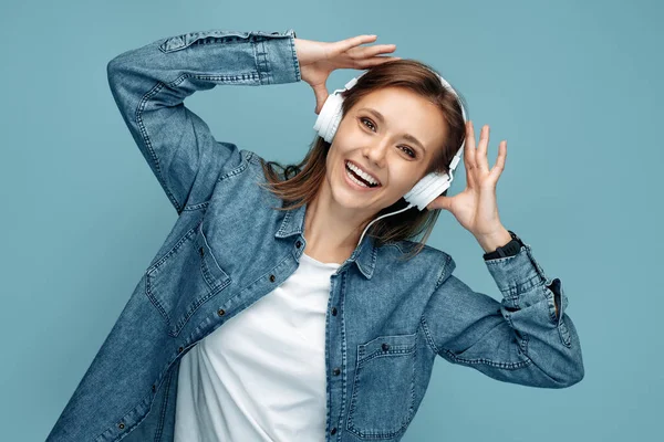 Positive young woman in jeans shirt listening music on headphone over blue background. — Stockfoto