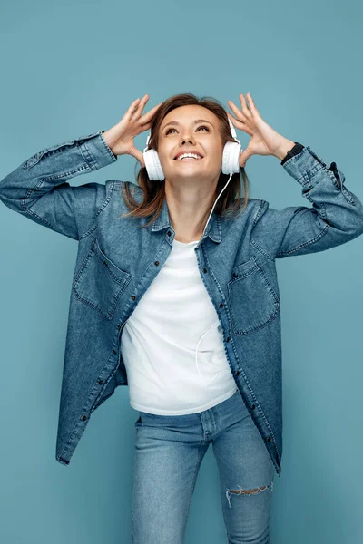 Unvorsichtige junge Frau in Jeanshemd, die über Kopfhörer vor blauem Hintergrund Musik hört. Freiheit. — Stockfoto