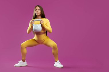 Tam uzunluk görüntüsü sarı spor kıyafetli esmer bir kadın mor arka planda kettlebell ile çömelme hareketleri yapıyor.